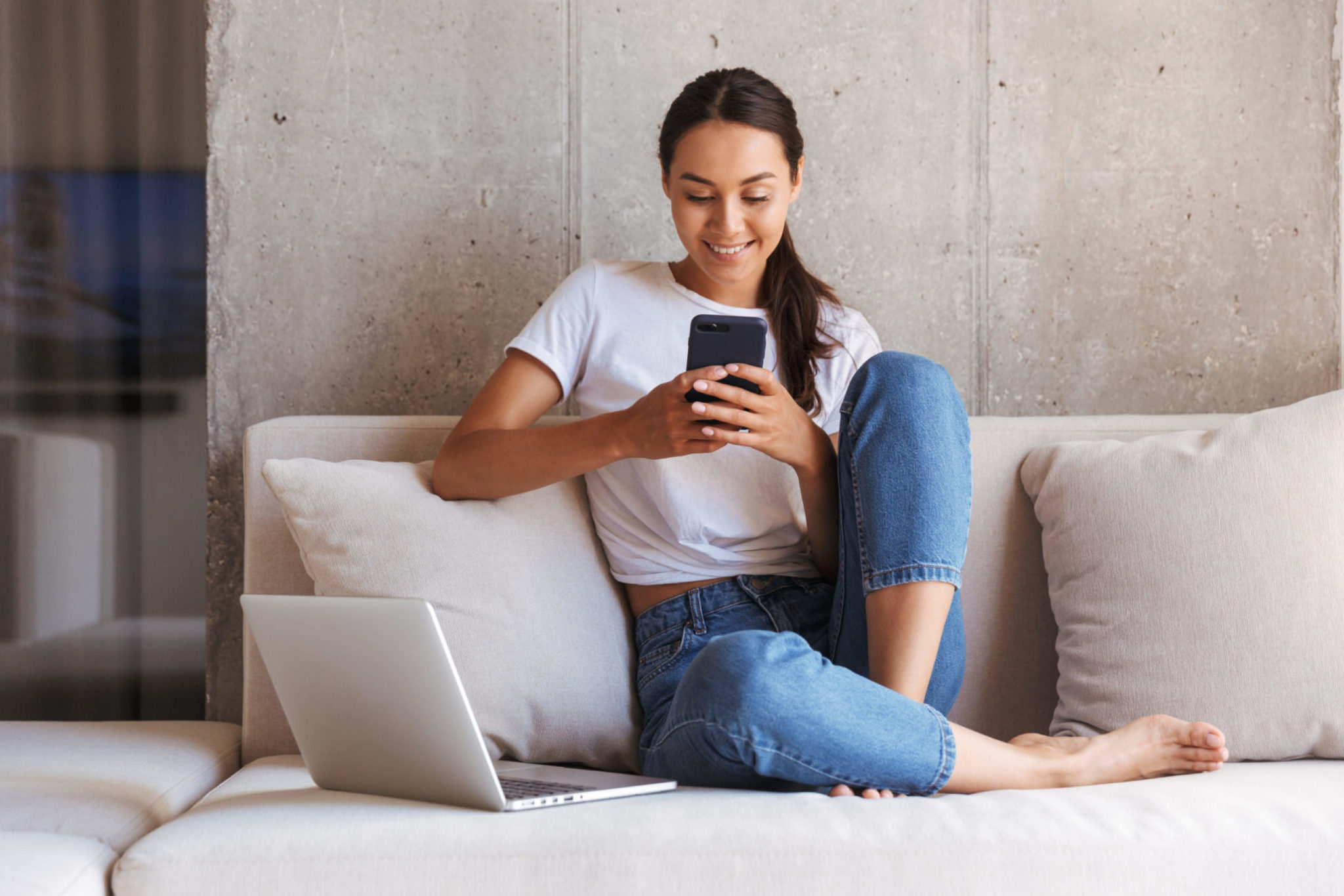Young adult using mobile phone on a couch with laptop opened beside them
